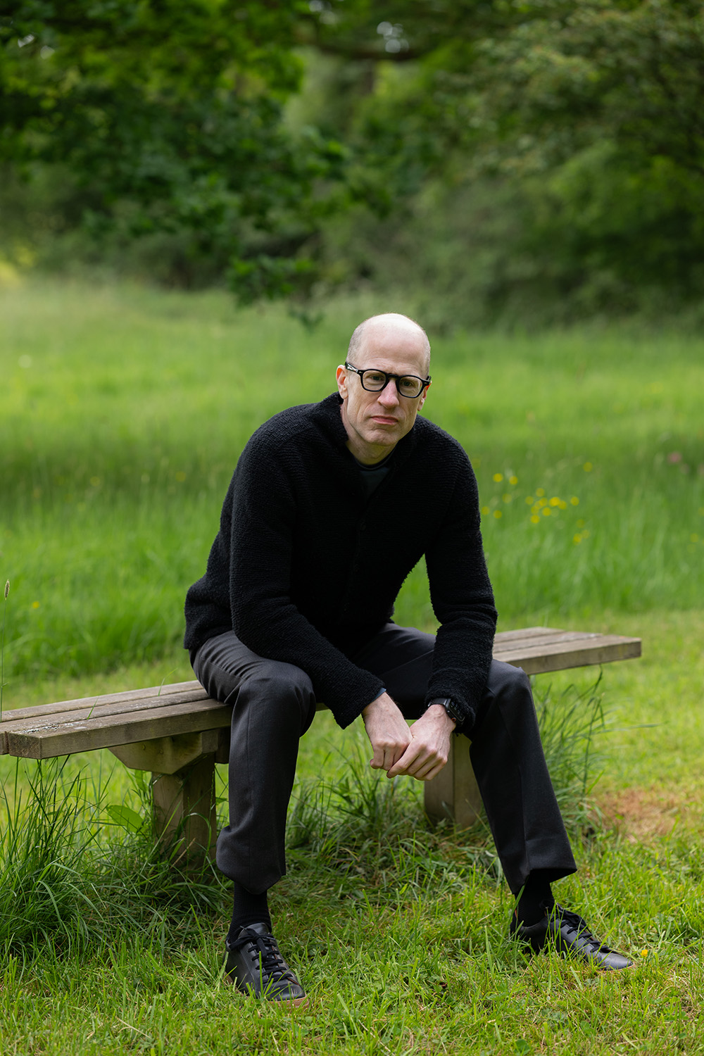 Outdoors portrait of Nick Bostrom sitting on a wooden bench in a forest meadow